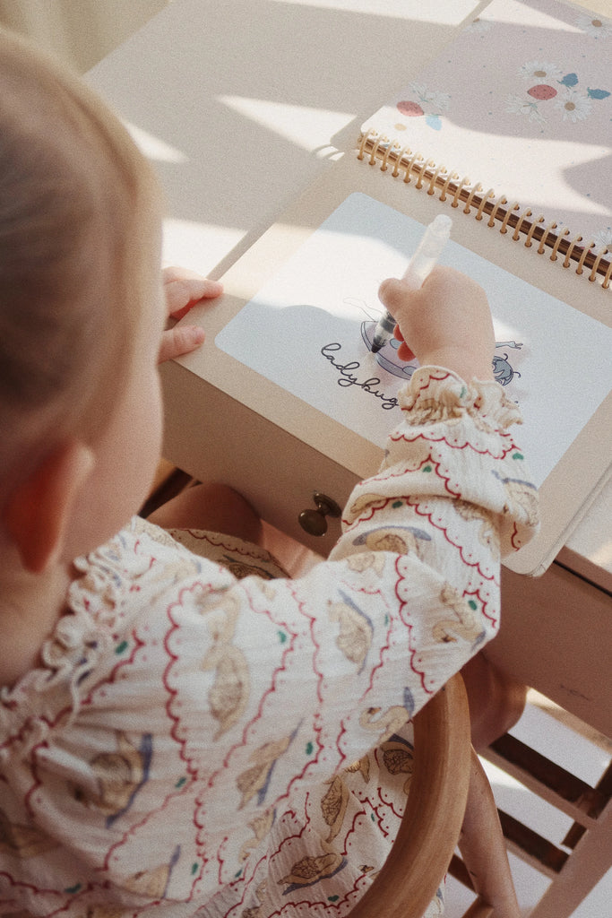 A child is drawing on a piece of paper in a spiral-bound sketchbook using a white crayon. The child is sitting at a light-colored desk and wearing a patterned, frilly outfit. The drawing features colors and shapes, but the specific details are not clear. Nearby lies My Magic Water Book - Off White, ready for more artistic fun.