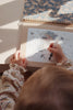 A child is coloring a picture of a unicorn riding the moon in a spiral-bound My Magic Water Book - Off White. The child is using a refillable water brush pen, with stars and hearts adorning the image around the unicorn. The child's hands and partial head are visible, and they are wearing a patterned top.