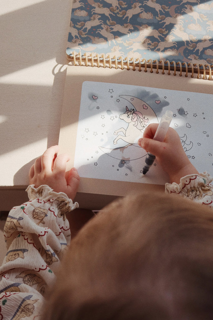 A child is coloring a picture of a unicorn riding the moon in a spiral-bound My Magic Water Book - Off White. The child is using a refillable water brush pen, with stars and hearts adorning the image around the unicorn. The child's hands and partial head are visible, and they are wearing a patterned top.