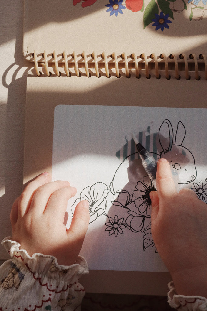 A child's hands are seen coloring in an illustrated page featuring a rabbit and flowers from the My Magic Water Book - Off White. The child is using a pen, with light and shadows beautifully cast on the reusable pages. The child is wearing a ruffled sleeve shirt with a delicate pattern.