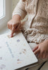 A child in a knitted sweater sits by a window, pointing at an illustrated storybook titled "My Magic Water Book - Sleet." This magic water coloring book features whimsical drawings of animals and stars, while the child’s other hand holds the edge of the book.