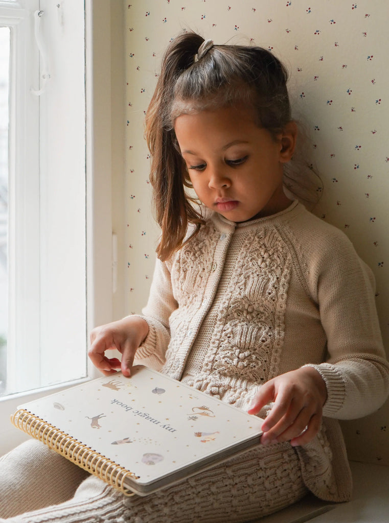 A young girl with long brown hair sits by a window and attentively reads a children's book. She is wearing a cozy, cream-colored knitted outfit and pointing to illustrations in what appears to be the My Magic Water Book - Sleet with reusable pages, set against light wallpaper featuring small, delicate patterns.