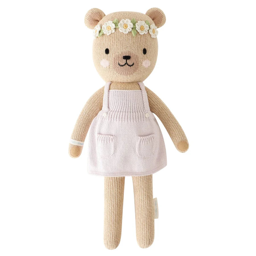 A hand-knit doll, the Cuddle + Kind Honey Bear is made from 100% cotton yarn and features a light pink apron-like dress with pockets. Its smiling face, rosy cheeks, and black nose are complemented by a floral headband of white flowers with yellow centers. The bear is filled with hypoallergenic polyfill for added softness.