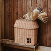 The Olli Ella Hanging Rattan Basket Hutch, shaped like a small house and featuring a charming window, rests on a wooden shelf against a wooden wall. The basket is filled with an arrangement of dried flowers and foliage, exuding a rustic and natural charm.