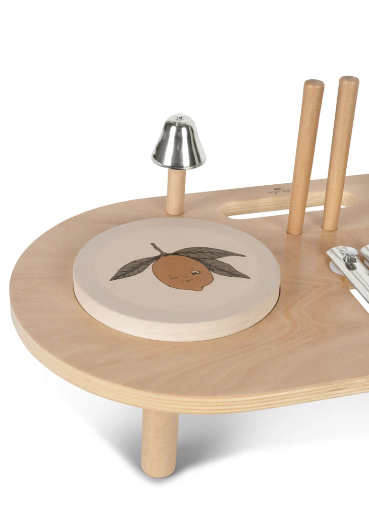 The Konges Sløjd - Wooden Music Board is crafted from FSC-certified plywood and features a printed lemon on a round cutting board, accompanied by a metal bell, utensils, and two wooden dowels on a smooth, light wood surface.