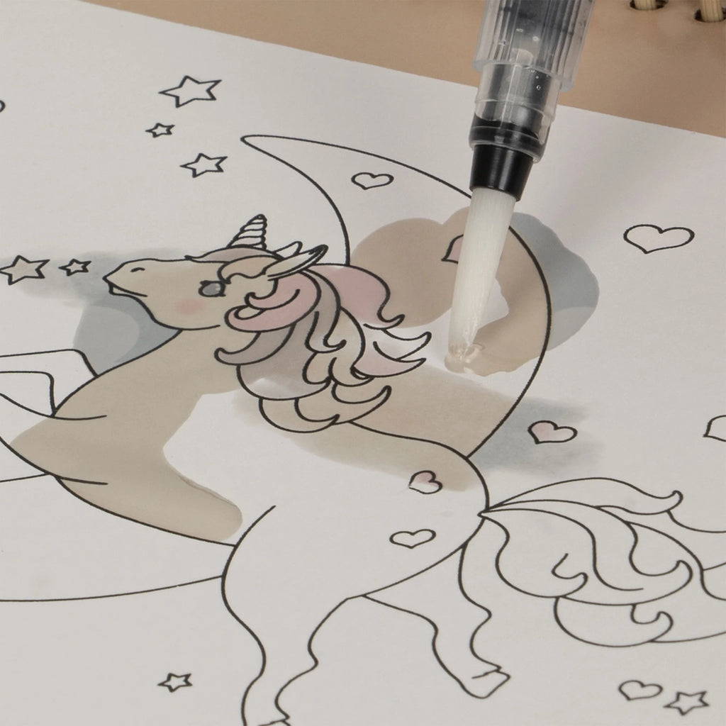 A page from the "My Magic Water Book - Off White" features a unicorn with a star-studded background and a crescent moon. The unicorn has flowing hair, a horn, and small hearts on its body. A refillable water brush pen brings the unicorn's mane to life with a soft pink hue on these reusable pages.