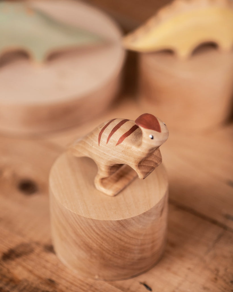 A handmade wooden Parasaurolophus dinosaur with handcrafted stripes stands atop a cylindrical wooden block on a wooden table, with a blurred background of more wooden items.