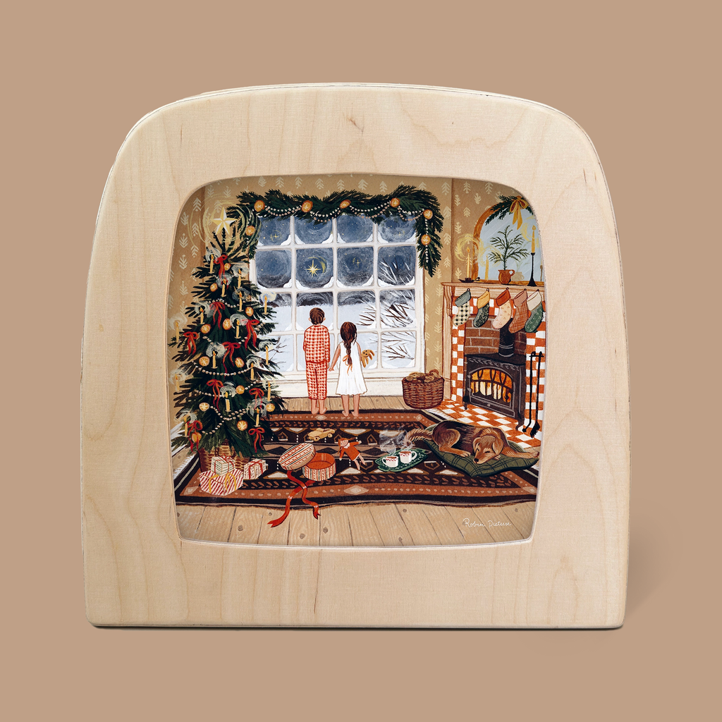 A cozy scene features the "Morning Star" silhouette, crafted for the Toverlux Lamp, depicting two figures standing by a window observing falling snow. Nearby, a decorated Christmas tree complements the setting with a fireplace, stockings, and gifts enhancing the warm, magical atmosphere.