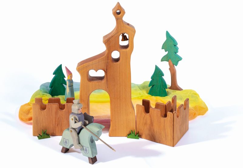 The Ostheimer Daughter of the Castle toy set includes a knight on horseback with a lance, a castle complete with walls and towers, a figure wearing a pointed hat, and background trees. Handcrafted from wood and featuring colorful fabric accents, this enchanting scene is ideal for fostering imaginative play.