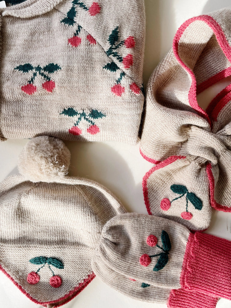 A collection of winter clothing including a Sonja Knit Mittens - Beige Melange with red and green cherry pattern, matching knit hat with a pom-pom, and coordinated socks on a white surface