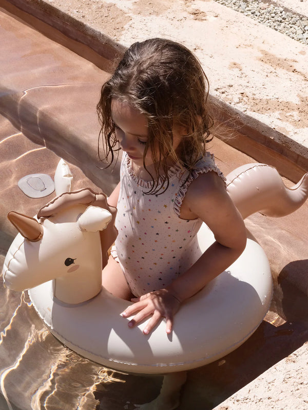 A young girl sits in a small pool on a sunny day, playing with an Inflatable Junior Swim Ring - Unicorn. She is wearing a pink polka dot swimsuit, and her focus is on the water.