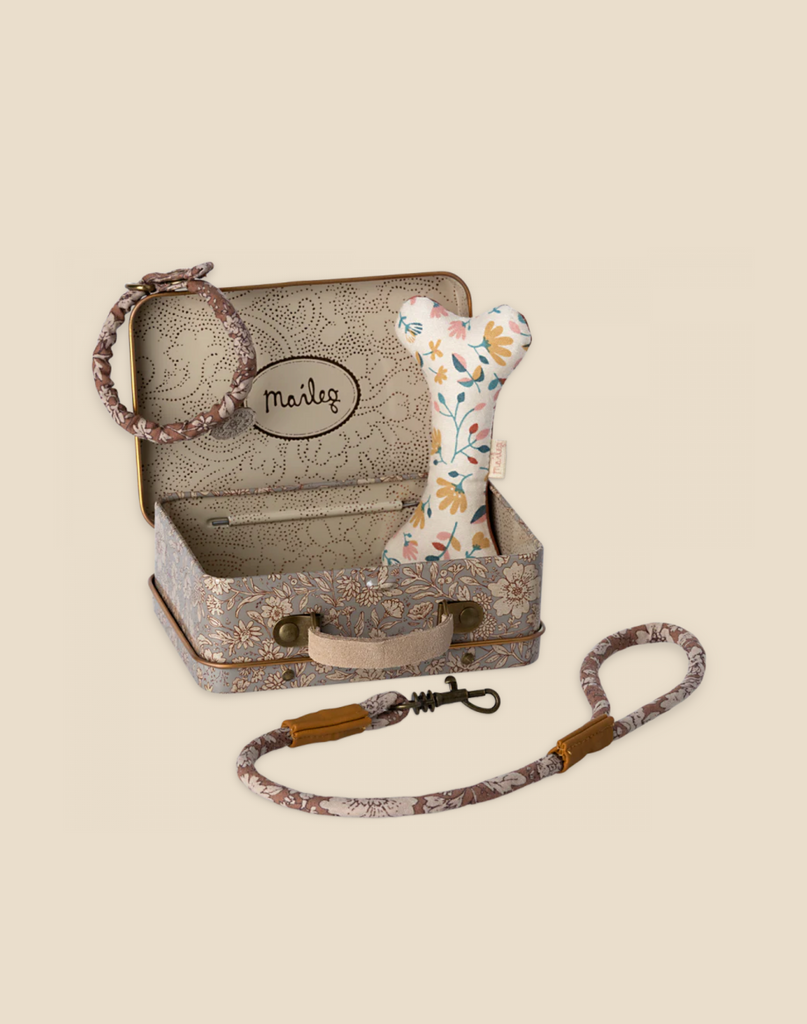 A decorative metal suitcase with floral patterns contains a plush dog, a floral print dog bone toy, a rope toy, and a matching collar and leash on a beige background. The word "Maileg Puppy Accessories - Rose" is printed on an oval inside the suitcase lid.