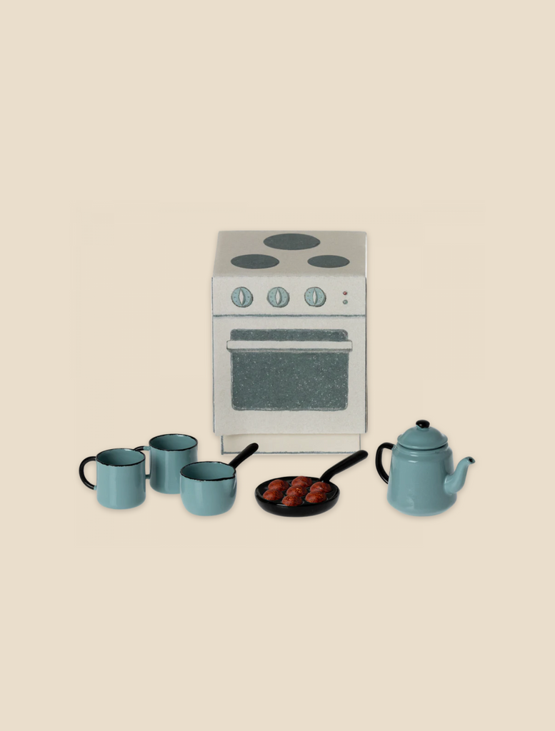 A miniature kitchen set featuring a white oven with silver knobs, a blue kettle, three blue mugs from Maileg Madam Blue’s Favorites, and a small frying pan with brown food items. The items are arranged on a light beige background, perfect for hosting the Mouse family's traditional Scandinavian treat.