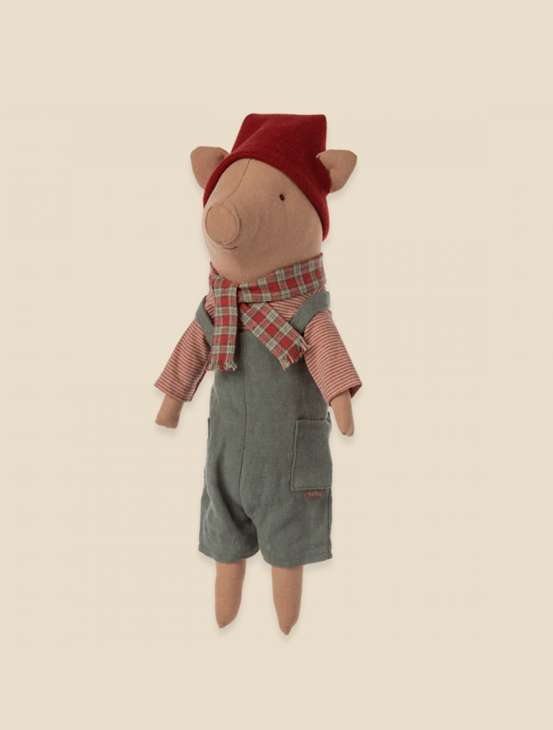 The Maileg Christmas Pig - Boy is a snuggly plush toy dressed in denim overalls and a red shirt, complemented by a red and green checkered scarf and a removable matching hat. Perfect as a holiday decoration, this charming pig features floppy ears and an embroidered snout, standing against a light beige background.