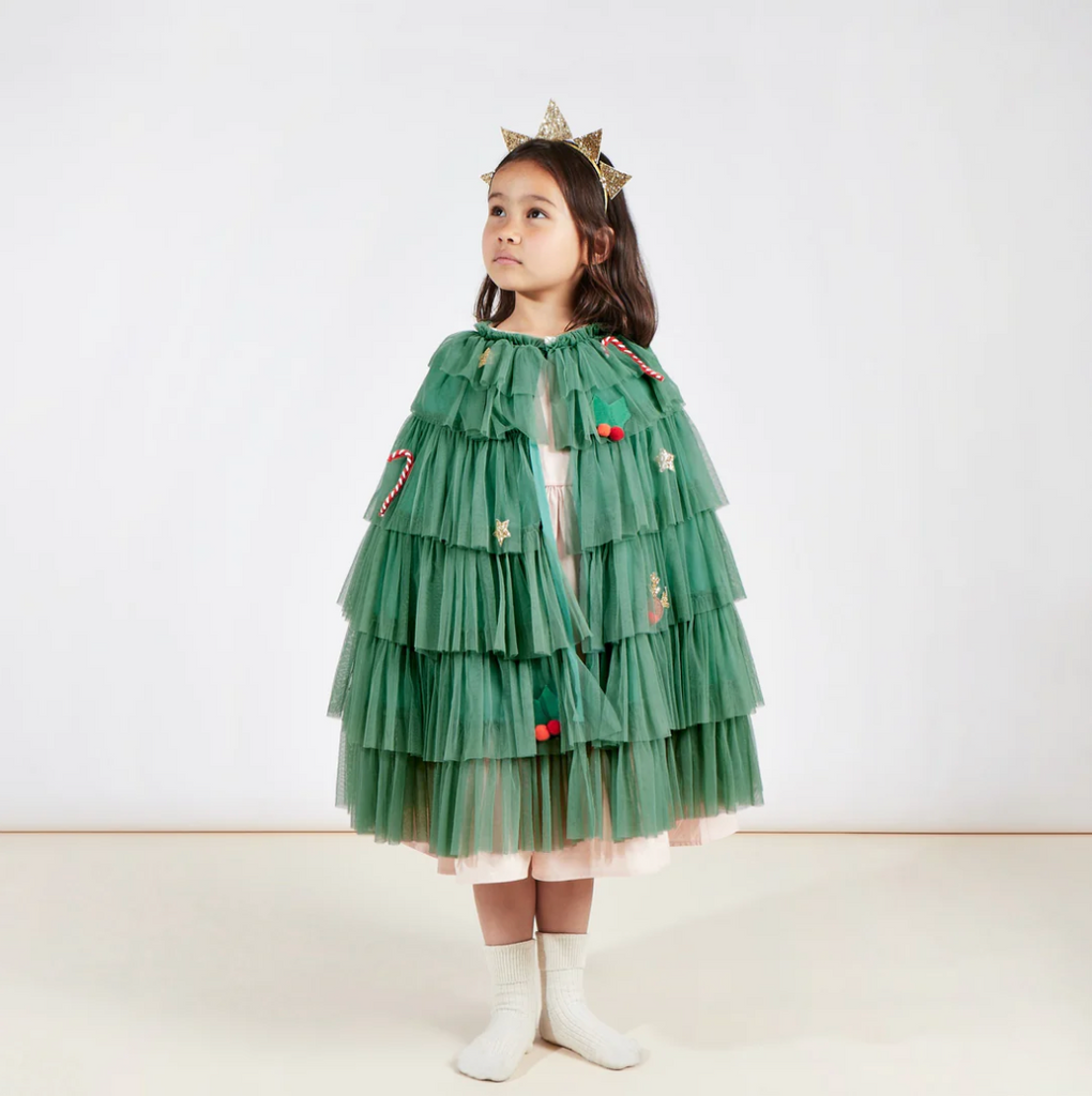 A young girl wearing a Meri Meri Tree Cape Costume adorned with red embellishments and a gold star headband stands against a plain background, looking to the side thoughtfully.