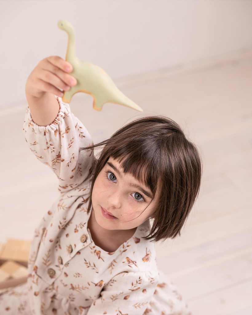 A young girl with short brown hair and a floral dress is looking up while holding a handmade Wooden Brachiosaurus Dinosaur above her head, sitting on a wooden floor.