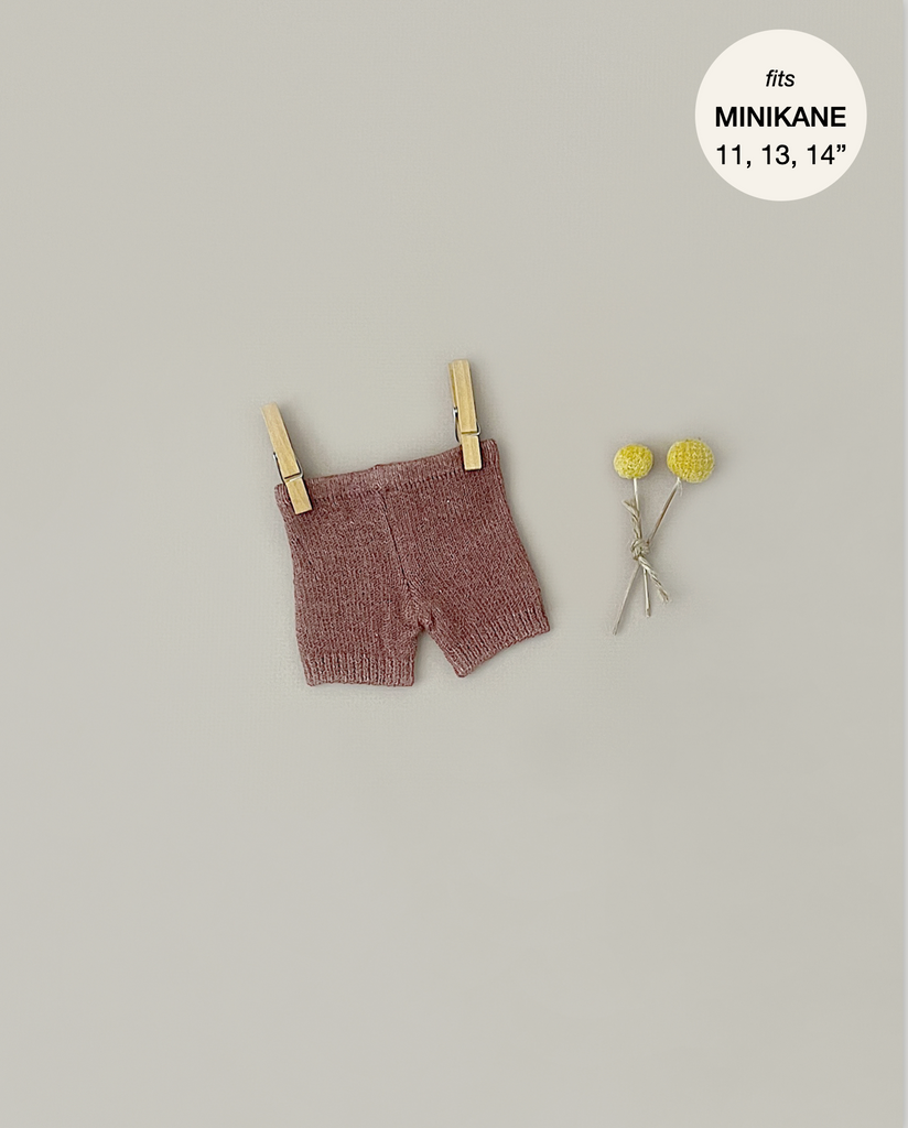 Minikane Doll Clothing | Vito Shorts in Marl Caramel Knit hang from two wooden clothespins against a light grey background. Beside the shorts, a small bouquet of three yellow pom-pom flowers adds a touch of charm. A round label in the top right corner reads "fits MINIKANE 11, 13, 14".