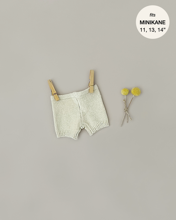 A pair of Minikane Doll Clothing | Vito Shorts in Cream Knit is clipped to a clothesline with two wooden clothespins. The background is plain beige. Two small yellow flowers are positioned on the right side of the shorts. Text in the top right reads, "fits MINIKANE 11, 13, 14” – perfect for Minikane Babies or Gordis dolls.