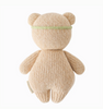 A beige, knitted Cuddle + Kind Baby Honey Bear, Ivory Floral with round ears and a green headband stands upright, facing away to showcase its back. This heirloom-quality bear is hand-knit from natural Peruvian cotton yarn, with the texture of the knit pattern visibly detailed.