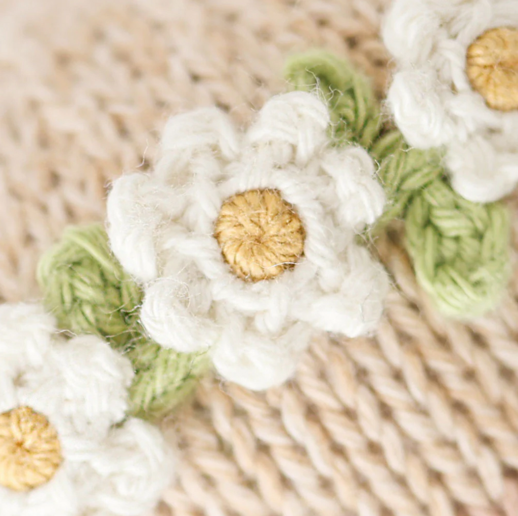 Close-up of a Cuddle + Kind Baby Honey Bear, Ivory Floral featuring white flowers with yellow centers and green leaves, crafted from natural Peruvian cotton yarn. Placed on a knitted beige background, the heirloom-quality texture and detail in the crochet work are clearly visible, highlighting the craftsmanship.