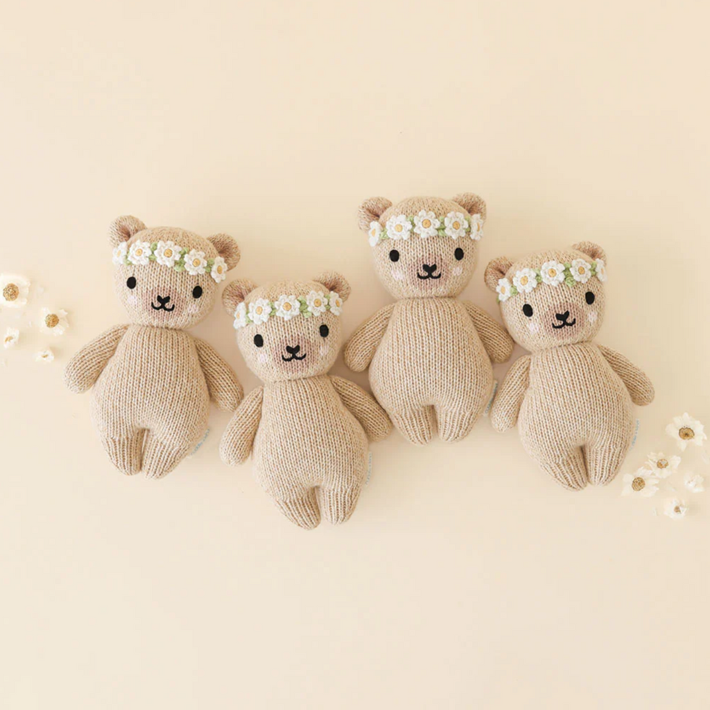 Four **Cuddle + Kind Baby Honey Bear, Ivory Floral**, hand-knit from natural Peruvian cotton yarn, wear floral crowns as they stand in a row against a beige background. Each bear features cute expressions with black button eyes and small smiles, while small white flowers are scattered around them.