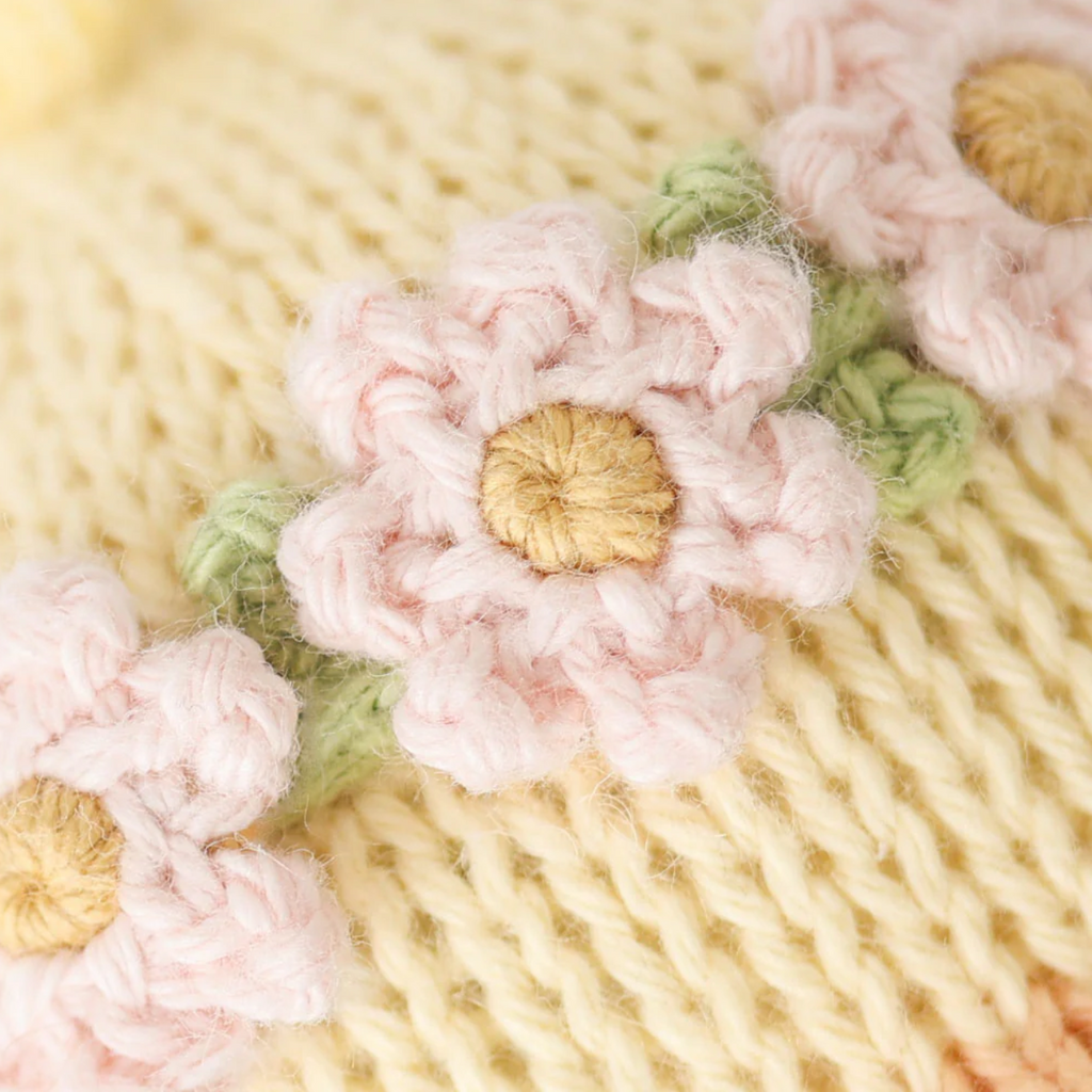 Close-up of an heirloom-quality knitted fabric featuring pastel-colored crocheted flowers. The background is a soft yellow knit, and the flowers are pale pink with green leaves, all crafted from luxurious Peruvian cotton yarn, creating a delicate and charming design. Cuddle + Kind Baby Duckling, Blush Floral