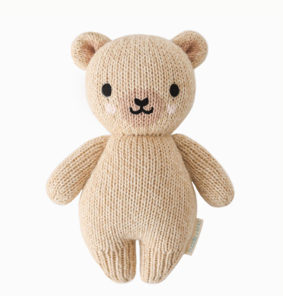 A Cuddle + Kind Baby Honey Bear made from natural Peruvian cotton yarn, featuring black eyes, a small black nose, and a smiling mouth. The collectible bear has round ears and is standing upright on a white background, embodying the charm of hand-knit baby animals.