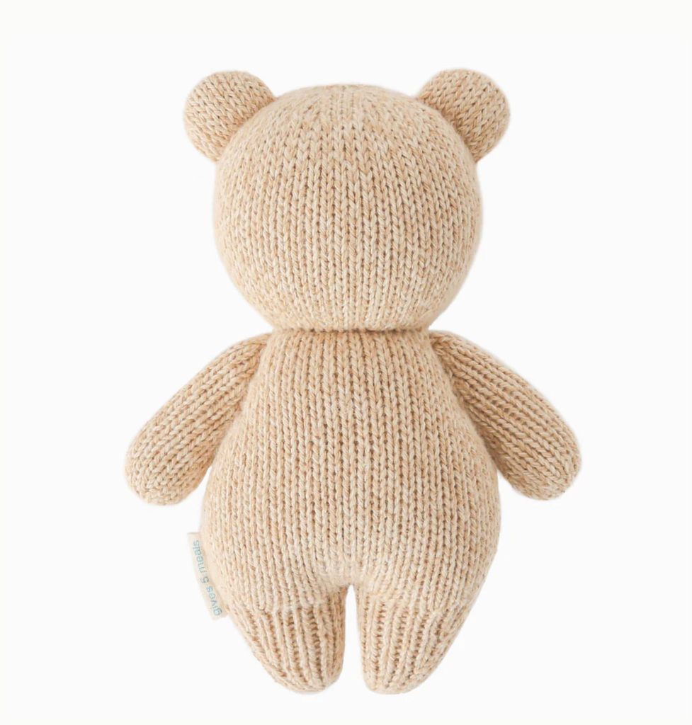 A beige knitted *Cuddle + Kind Baby Honey Bear* made from natural Peruvian cotton yarn, showcasing a simple, minimalist design. The bear is shown from the back, featuring rounded ears, short arms, and legs. The texture of the knit is visible, giving it a cozy and handmade appearance perfect for any collectible set.