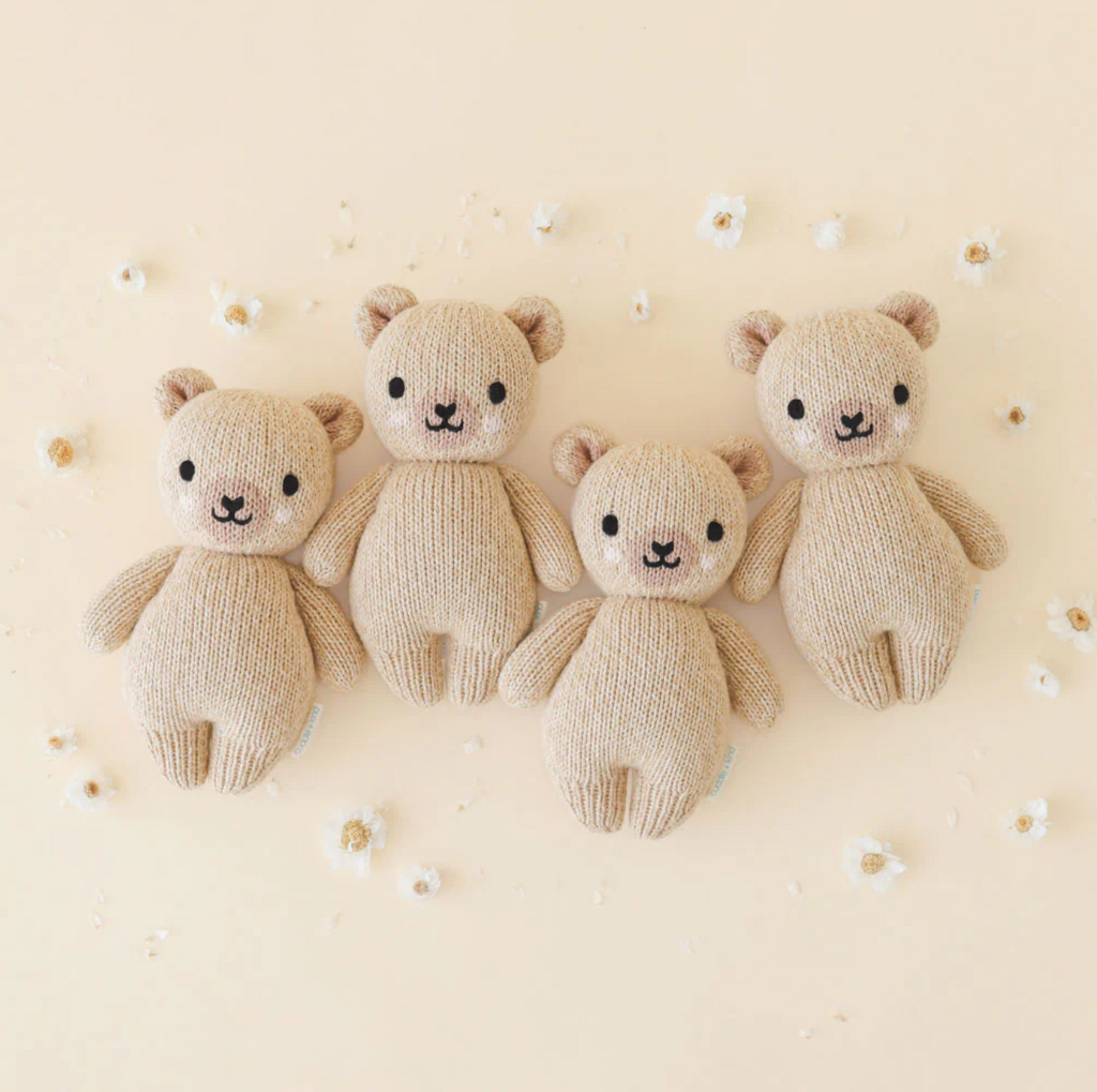 Four Cuddle + Kind Baby Honey Bear, hand-knit baby animals made from natural Peruvian cotton yarn, are arranged in a row on a pastel beige background. Each bear has a light brown color with black eyes, a small black nose, and small black threads for mouths. Small white floral decorations are scattered around the collectible bears.