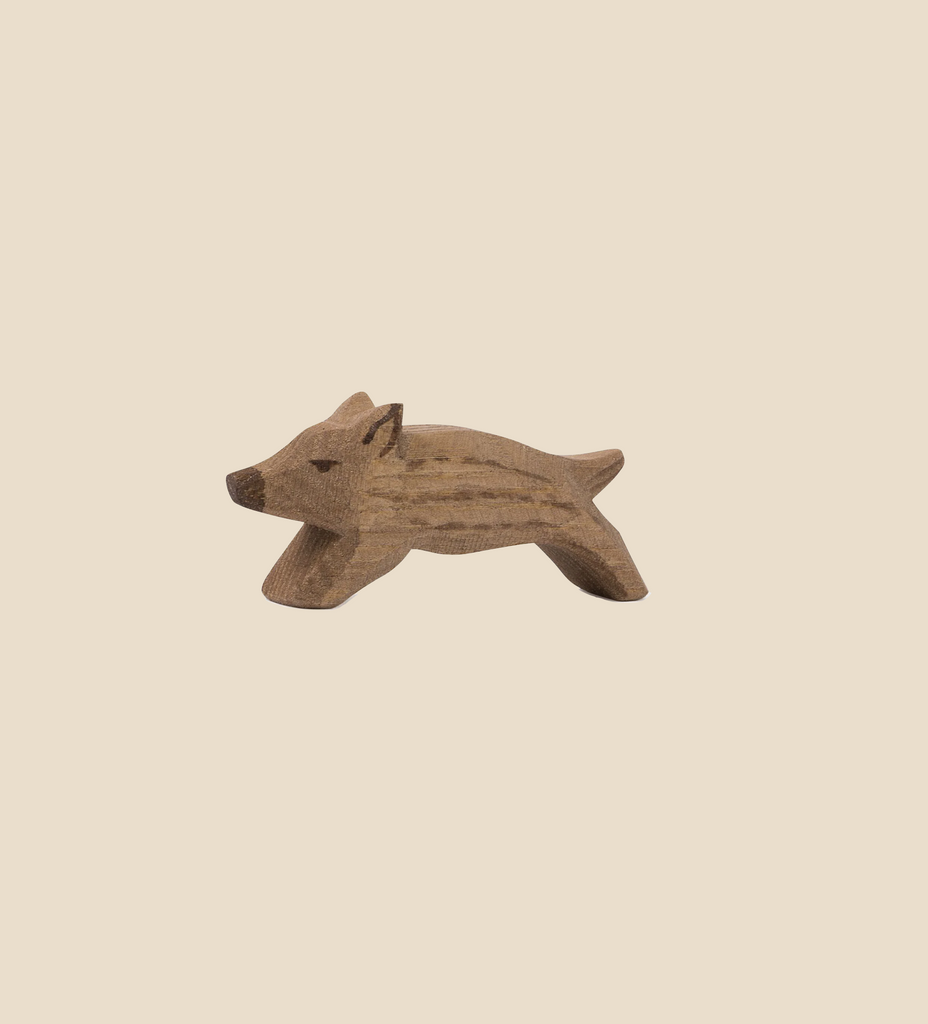 A small, light brown wooden figurine of a wild boar in a playful stance, handcrafted by Ostheimer, placed on a beige background.