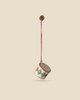 A small, decorative Maileg Metal ornament, Small drum - Classic green hangs from the ceiling, featuring a red and green diamond pattern and is suspended by a red and white twisted cord against a plain beige background, reminiscent of sparkling Maileg ornaments on a Christmas tree.