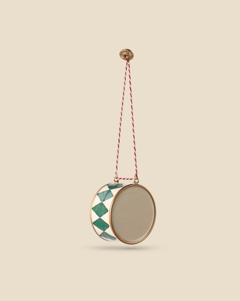 The Maileg Metal Ornament, Large Drum in Dark Green features a circular design with an elegant gold frame and handpainted green and white diamond patterns on the sides. It hangs from a stylish red and white twisted rope, which is secured by a brass wall hook, creating a stunning display against a light beige background.