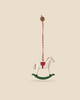 The Maileg Metal Ornament, Rocking Horse with a red saddle and striped hanging string sways gently against a beige background. This charming piece, featuring handpainted details and a simple design, rests on a green base.