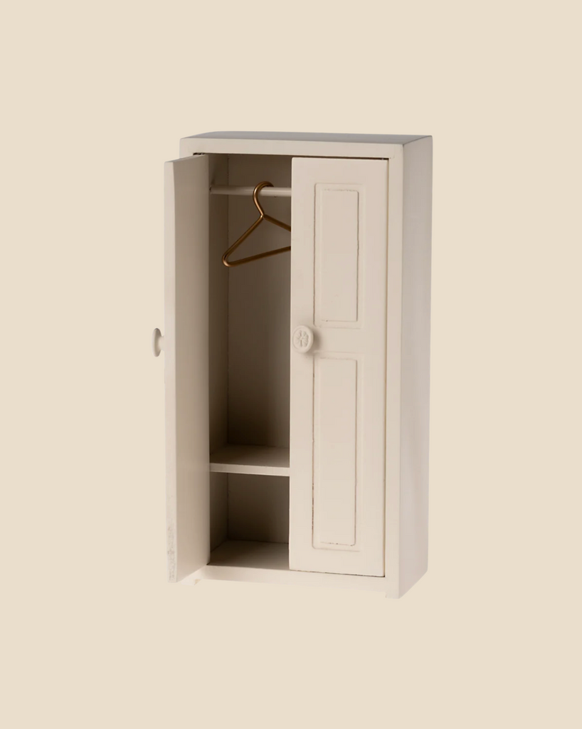 A petite, cream-colored Maileg Mouse Closet stands with one door slightly ajar, showcasing an empty interior featuring a golden hanger and two shelves. Set against a plain beige background, it's as though a tiny mouse family could consider this cozy closet their own. (Ships in approximately one week.)