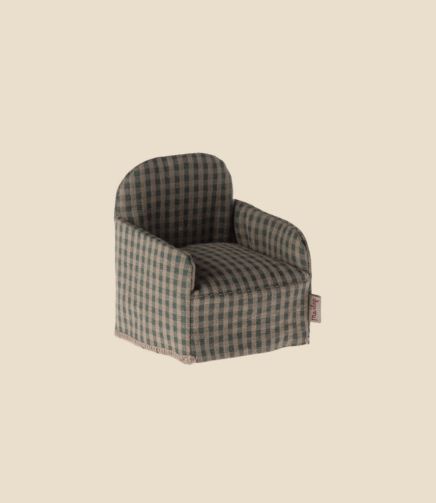 The Maileg Chair, Mouse - Green Checker features a small armchair design with a green and brown checkered pattern, set against a light beige backdrop reminiscent of a cozy gingerbread house. It has a soft, rounded back and arms, complemented by a small fabric tag on the side.