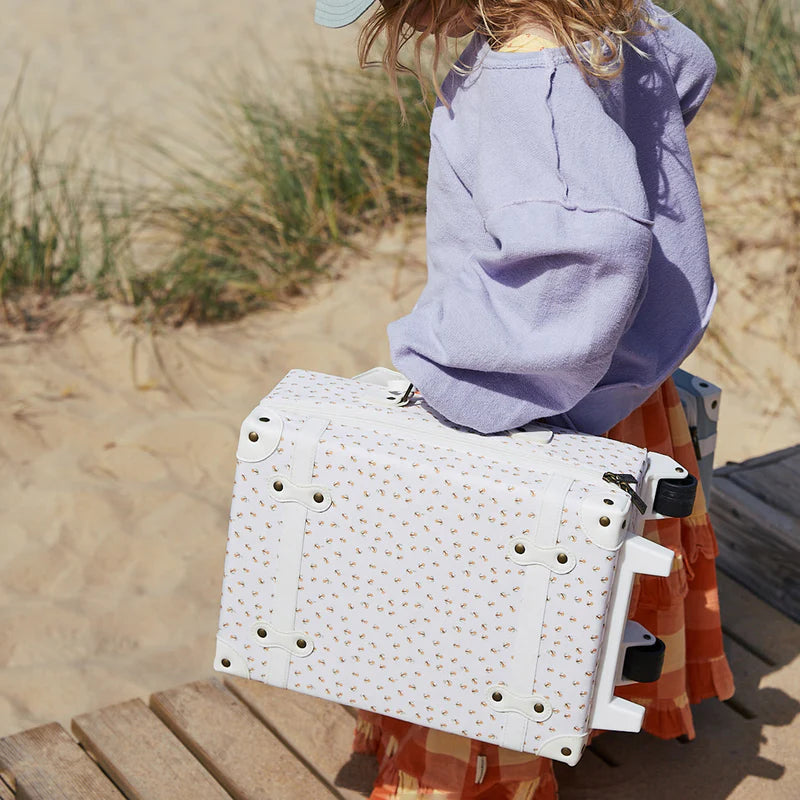 A young girl in a purple top and patterned skirt carries an Olli Ella See-Ya Suitcase - Leafed Mushroom, walking along a wooden boardwalk by a sandy beach.