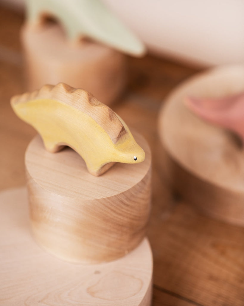 A small, Handmade Wooded Stegosaurus Dinosaur, painted yellow with non-toxic paint and minimalistic details, rests on a rounded wooden platform. Other handcrafted wooden toys in muted colors are slightly blurred in the background. The surface they occupy appears to be a wooden table.