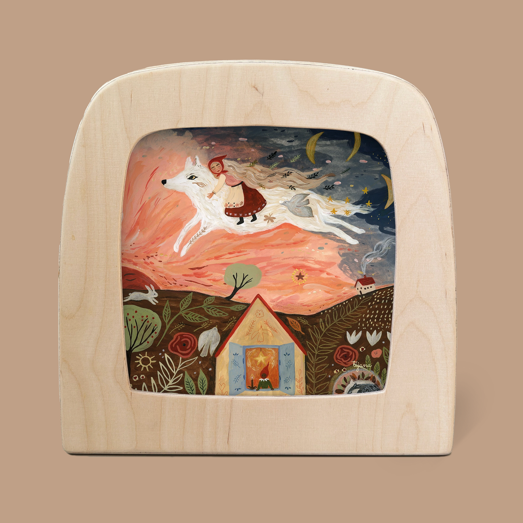 The silhouette-only design, "Girl and the Wolf," for the Toverlux Lamp by Tijana Draws showcases a whimsical scene of a girl riding a white wolf through a starry sky with celestial elements. Below, there's a cozy house amid flowers and foliage, all elegantly framed by a light wooden border reminiscent of enchanting Magic Silhouettes.