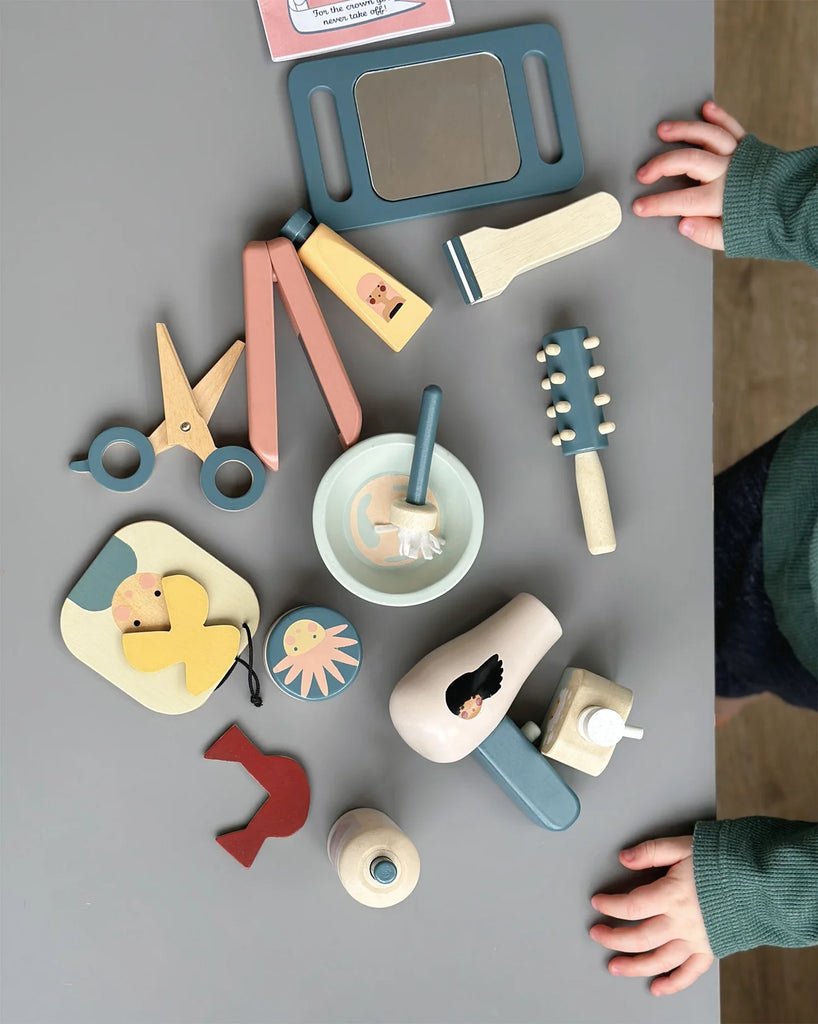 A child's hands reaching towards a collection of colorful wooden toys and craft supplies arranged neatly in a Hair Salon. Items include a drawing pad, scissors, a paintbrush, and decorative shapes.