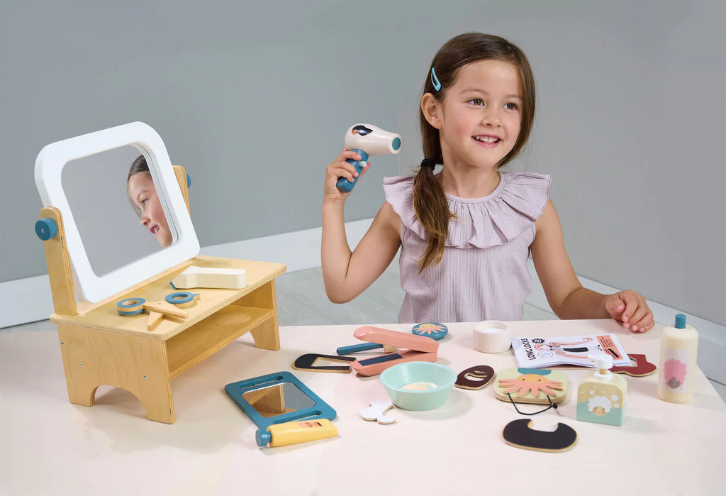 A young girl happily plays with a Little Tikes My First Hair Salon vanity set, holding a play hairdryer, with various accessories like brushes, mirrors, and pretend makeup on the table.