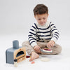 A young child in a striped sweater engages in pretend play with a Wooden Make Me A Pizza Set on a white background, slicing a toy eggplant with a toy knife.