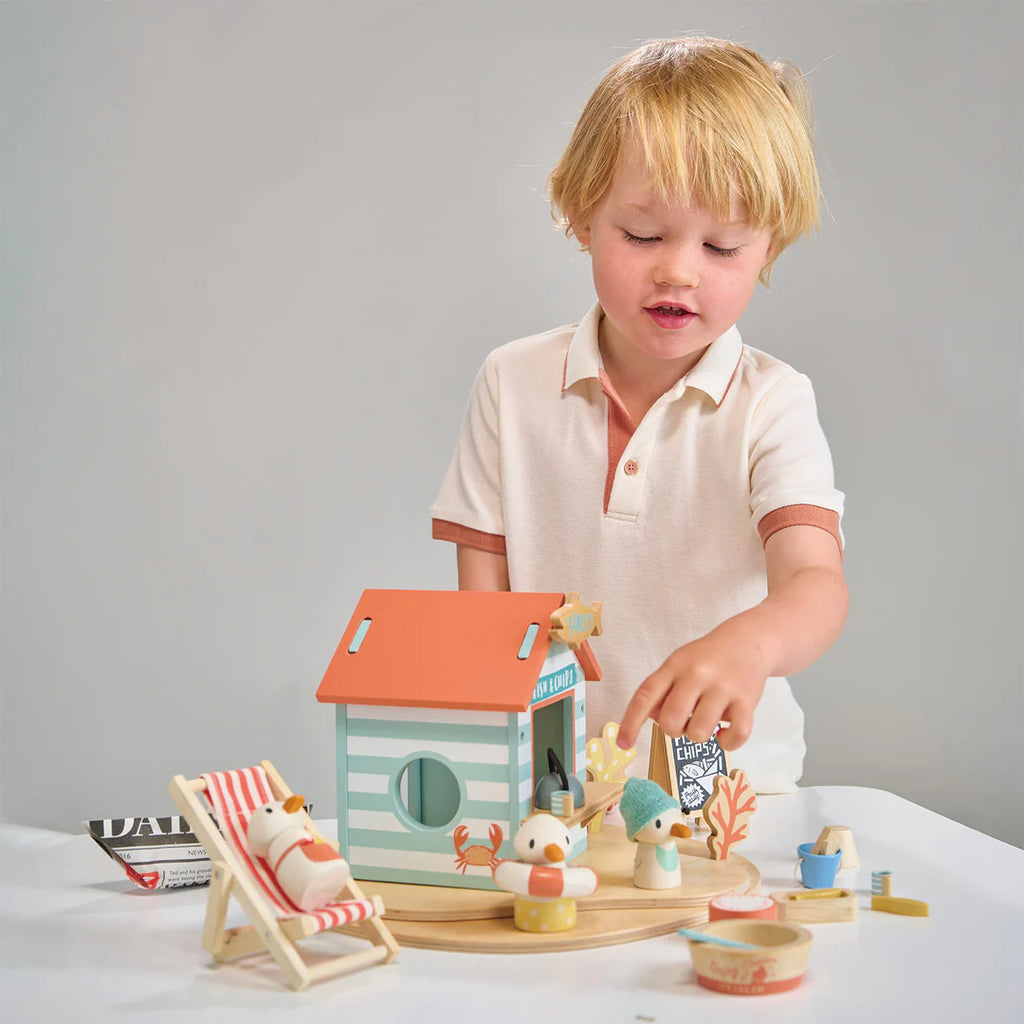 A young child engages in imaginative play with Sandy's Beach Hut and various wooden toys made of sustainable wood on a table, displaying curiosity and deep engagement.