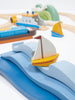 A close-up of a colorful children's wooden toy set featuring boats and waves. In the foreground, a small sailboat with a yellow and white design glides over blue solid wood waves. In the background, additional boats and sea creatures are partially visible, encouraging open-ended play with the Blue Water and Sail Boat Set.