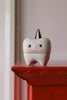 A small ceramic tooth-shaped container with a smiling face and closed eyes sits on a red shelf. This adorable Konges Sløjd Stone Tooth Box has a white base and a pointy cap on top. Perfect for holding a child’s first teeth, the background is light-colored and slightly out of focus.