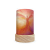 The Toverlux Lantern Shade Set - Holidays features a cylindrical art piece on a wooden base, crafted from translucent materials, showcasing an abstract figure with outstretched wings in warm reds, oranges, and yellows that produce a glowing effect reminiscent of lantern silhouettes. It ships in approximately one week.