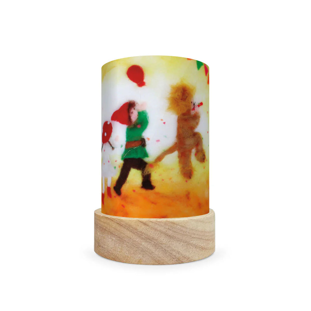 The Toverlux Lantern Shade - Seasons features a cylindrical design with a vibrant illustration of a child in a green outfit walking beside a creature holding a red balloon. Its base is made from light-colored wood, and the bright background emits an enchanting glow reminiscent of magic shades.