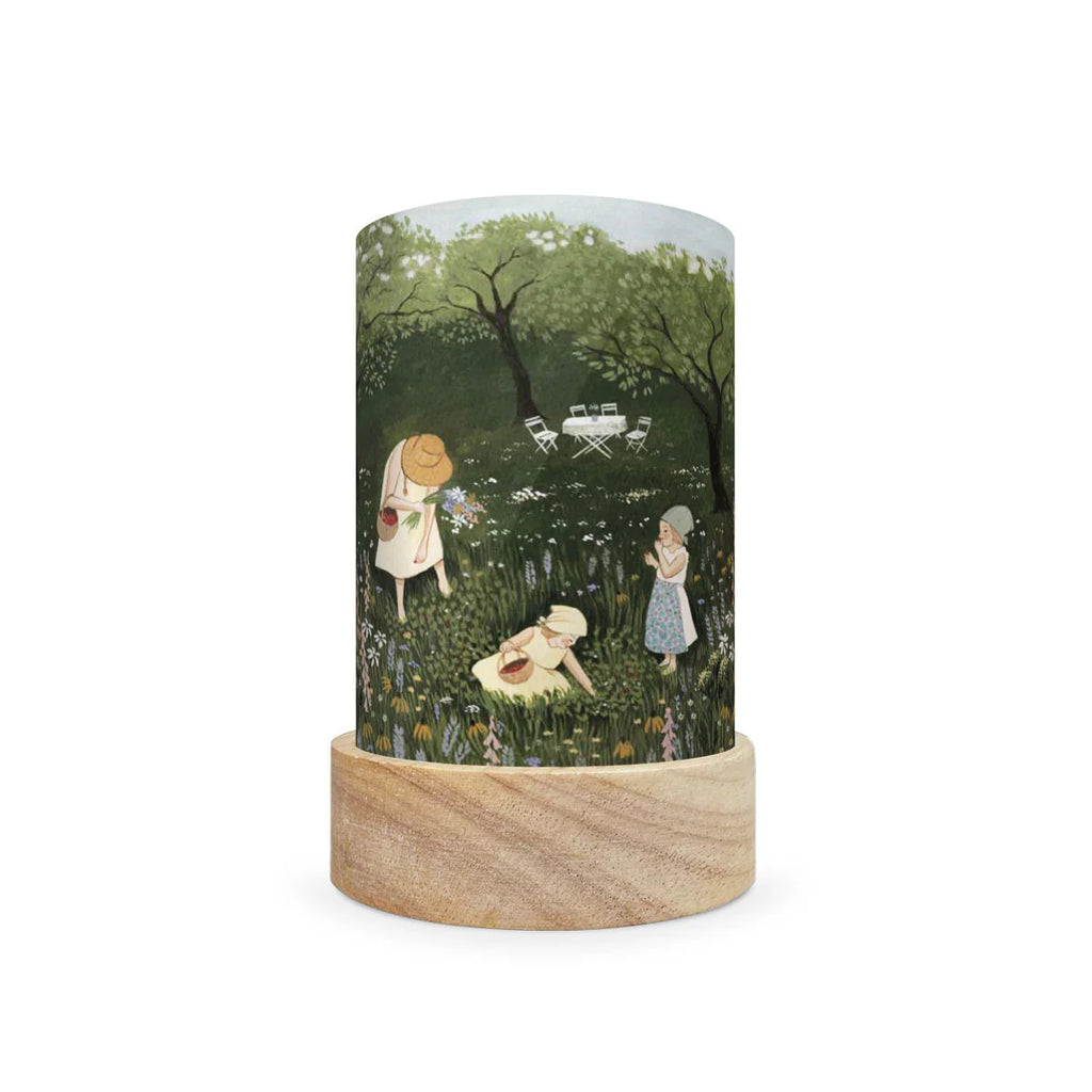 Introducing the Toverlux Lantern Shade Set by Robin Pieterse, a captivating cylindrical lamp featuring a delightful garden scene. It artfully depicts two people in hats and a child picking flowers among trees, accompanied by a small table and chairs. This enchanting piece, complete with its wooden base, brings magical silhouettes to life and ships in approximately one week.