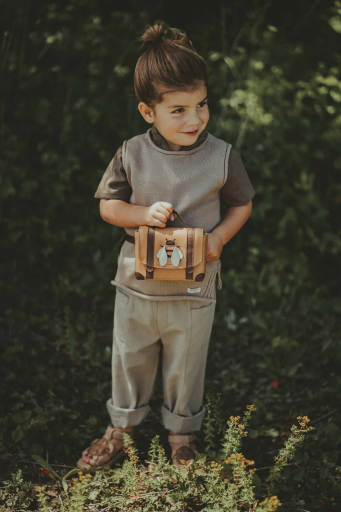 A young child holding a Donsje Trychel Bum Bag - Bee, smiling in a sunny garden surrounded by greenery and flowers. The child is dressed in casual summer clothing, sandals, and a waist bag with an adjustable leather strap