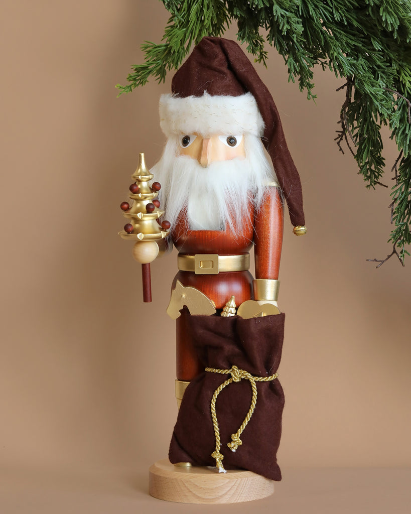 This limited-edition Christian Ulbricht Collectible Wooden Nutcracker - Natural and Gold Santa showcases a white-bearded Santa wearing a brown hat and holding a decorative golden tree. It is accompanied by a brown sack tied with a gold rope, while greenery elegantly drapes from the top right corner.