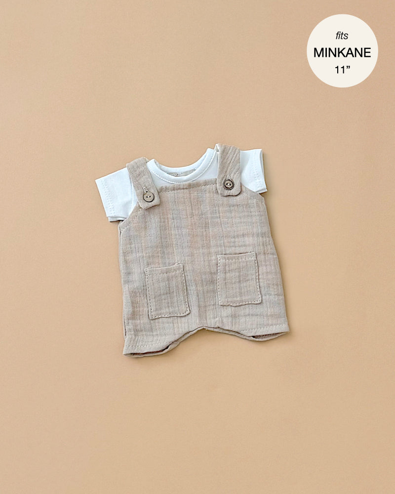 The Minikane Doll Clothing set includes Antonin Pebble Cotton Gauze Overalls and a White Jersey T-Shirt, designed specifically for an 11-inch baby doll. The beige overalls feature two pockets, making it a charming outfit set against a solid light brown background. This ensemble is not only stylish but also conveniently hand washable for easy care.